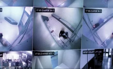 9 camera feeds on the one computer screen, showing blue-hued images looking down at bare cells. A blurred figure is curled on the floor of one cell image.