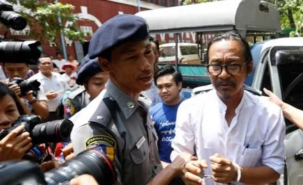 A man in handcuffs and led by police officers, with a media scrum around him
