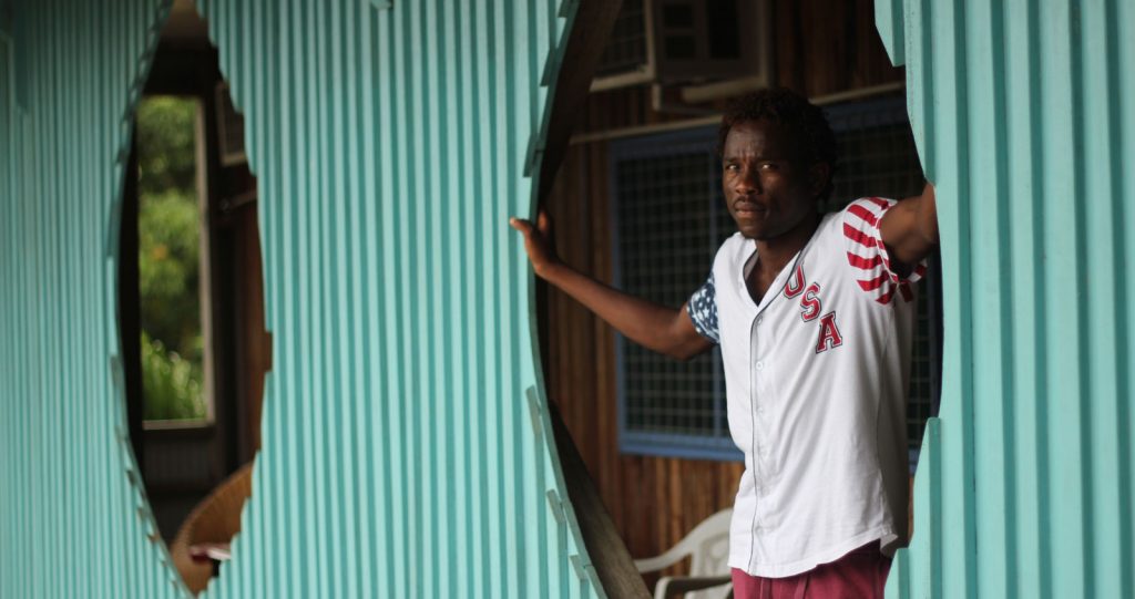 A man looks out a doorway of a wall made of teal green corrugated iron