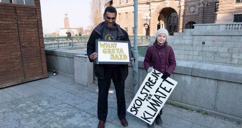 A teenage girl with a beanie and two long plaits holds a big handmade sign with words in Swedish: Skolstrejk for Climatet. Next to her, a dark-skinned man holds a handmade sign saying: What Greta said.