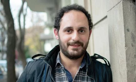 A young man leaning against a wall in a jacket, smiling kindly