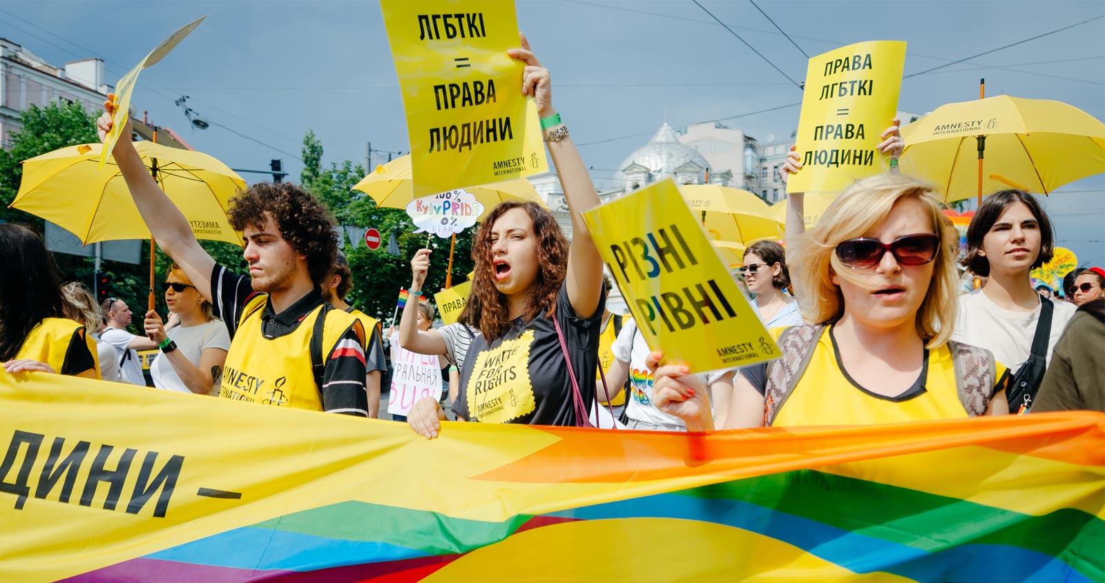 A rainbow coloured parade of people with banners and placards, marching on a sunny day.
