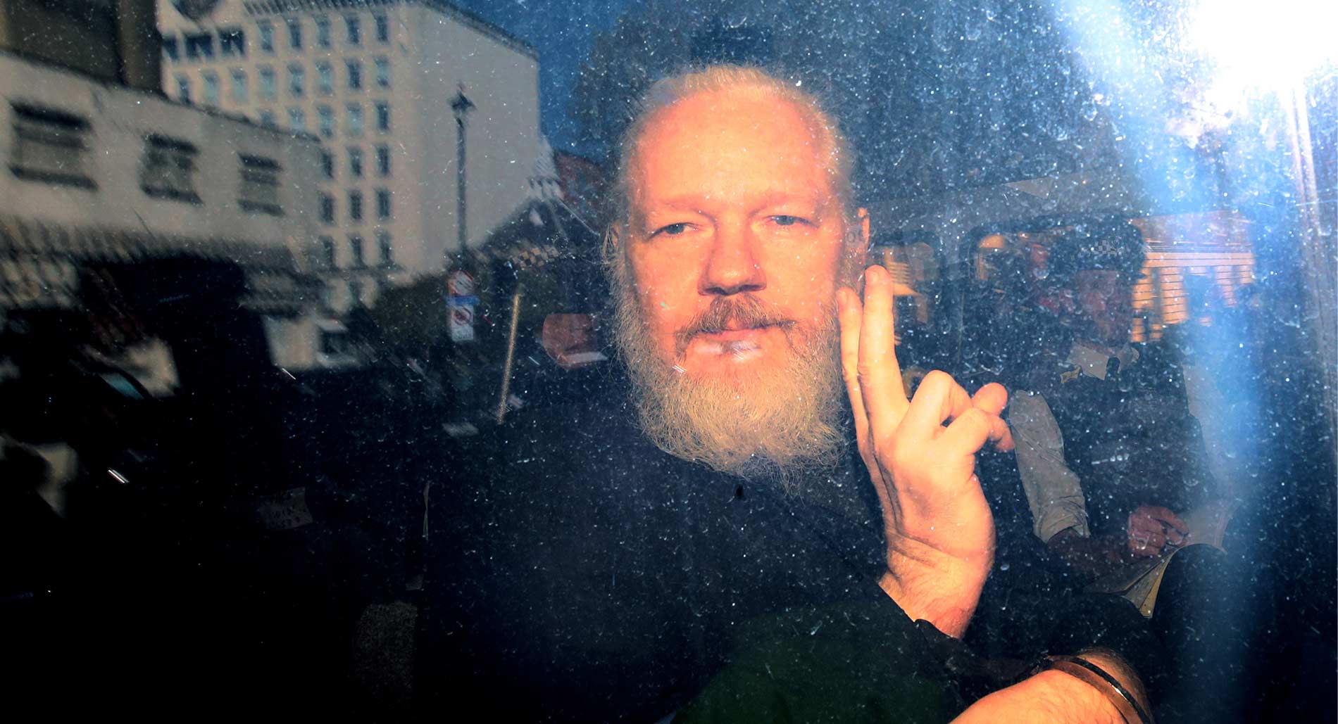 Julian Assange, a man with a white beard, raises his hand in a peace gesture with media and police around him. Twilight evening, bright flash of light to the right.