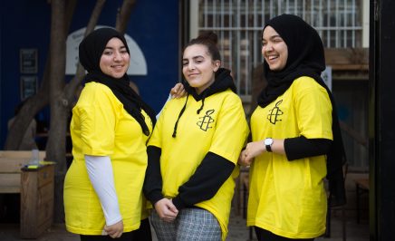 3 girls wearing bright yellow t-shirts, 2 with the hijab, smile in front of a tree and a dark blue wall. They are taking part in Amnesty France's Write for Rights event in Marseille.