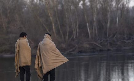Two refugees wrapped in blankets overlook a lake.