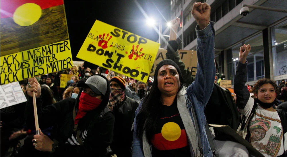 Protestors chant and bend down on their knees. They hold handmade red, yellow and black signs saying 'stop don't shoot' and Black Lives Matter. Woman in front raises her fist and is wearing an Aboriginal flag tshirt.