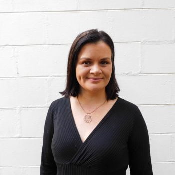 Portrait of Indigenous woman and activist Bianca Hunt. She stands in front of a white brick wall, smiling. She's wearing a black dress and pendant necklace.