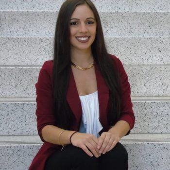 Portrait of Erika Rodriguez, refugee and women's rights activist. Erika is sitting on white steps, smiling. She's wearing a white top, black pants and maroon blazer.