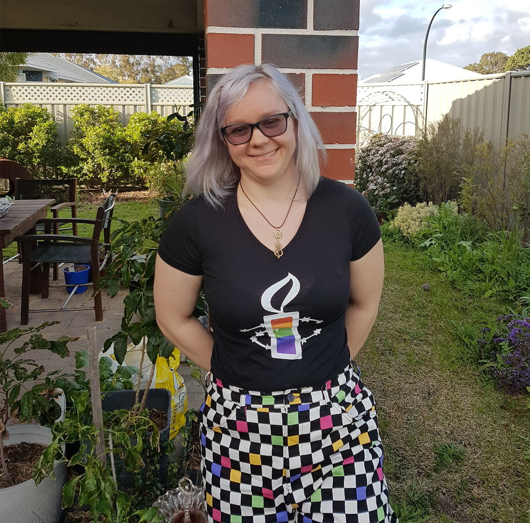 Image of LGBTIQ activist Hannah Wahlsten, smiling at standing in her yard. She's wearing rainbow-coloured checkered pants, an Amnesty tshirt with a rainbow candle, and black glasses.