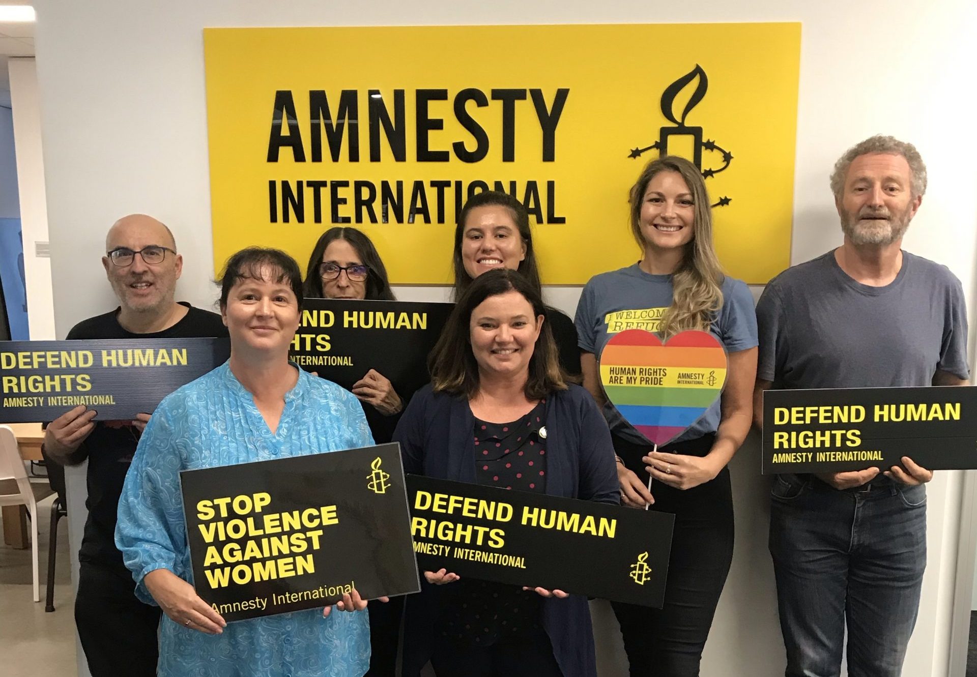 7 people holding signs saying Defend Human Rights standing in front of a yellow Amnesty International Sign