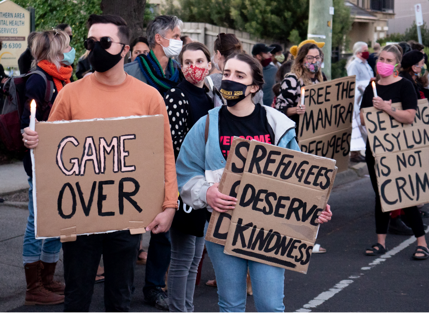 activists standing at a rally, wearing masks, holding cardboard signs reading "Game Over" "Refugees Deserve Kindness"
