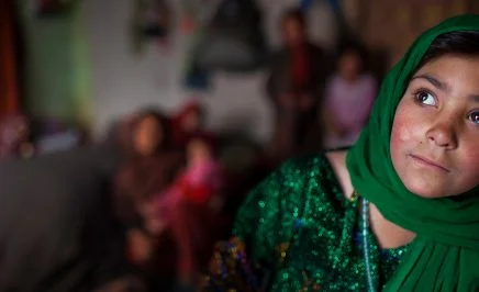 An image of a woman wearing a green hijab looks away from the camera. The background is distorted, but she is in a room with other people.