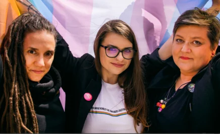 Elżbieta, Anna and Joanna stand in front of an LGBTQIA+ flag