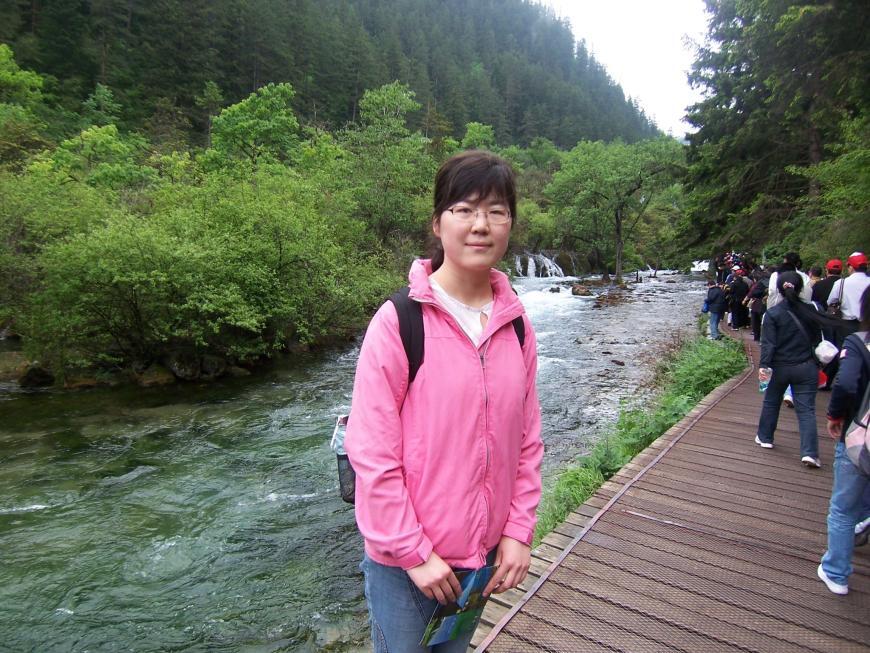image of women wearing pink jacket smiling into the camera in front of a riverside along a forest