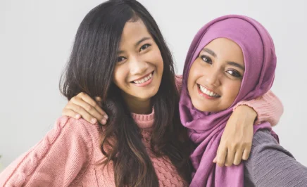 Two young women of colour, smiling. The girl on the left has long dark hair and has her arm around the girl on the right, who is wearing a purple hijab.