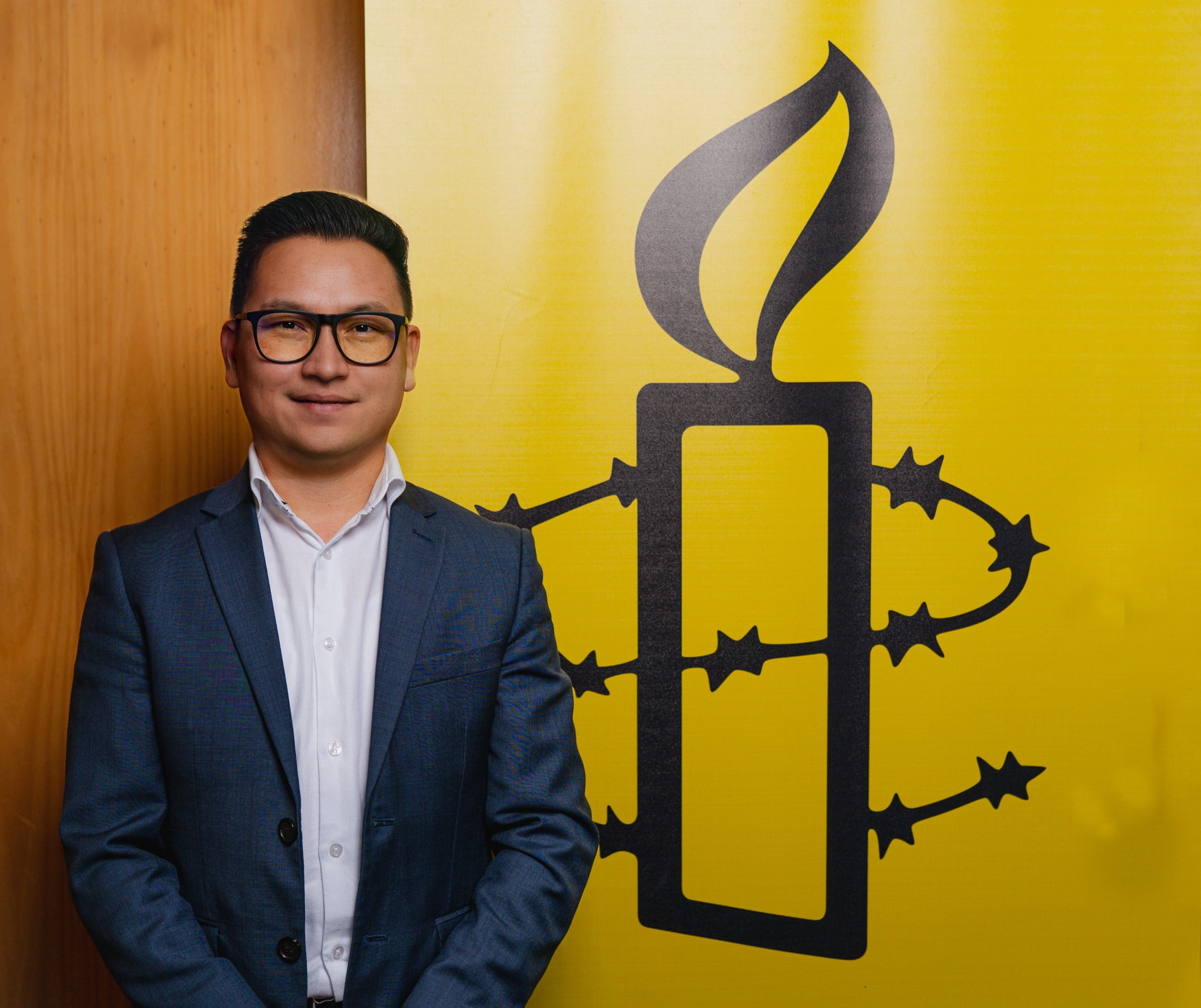 Amnesty International Australia Refugee Rights Campaigner Zaki Haidari pictured next to an Amnesty logo of a candle surrounded by barbed wire on a yellow background.